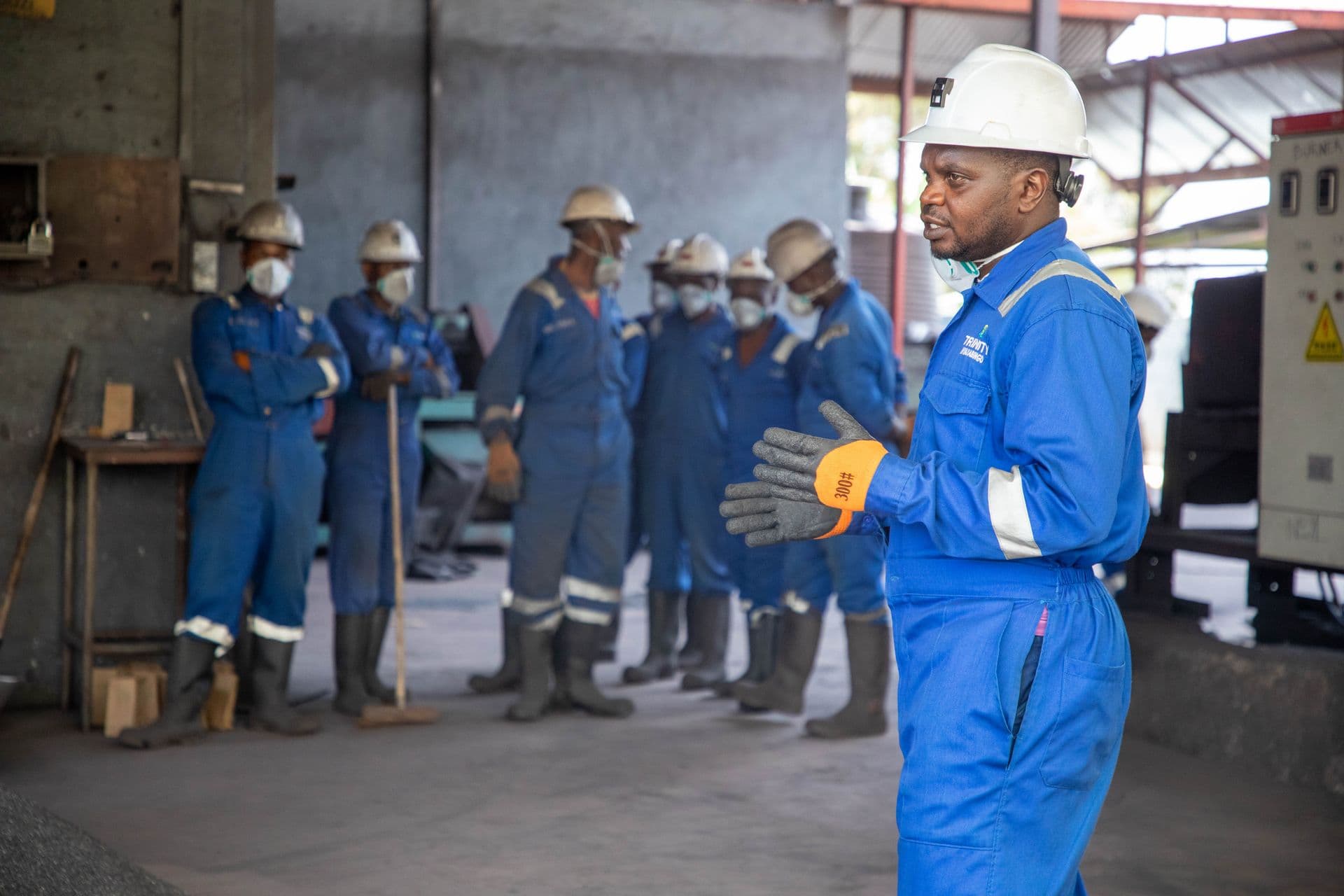 A worker in blue protective clothing and a white hard hat addressing colleagues in an industrial workshop.